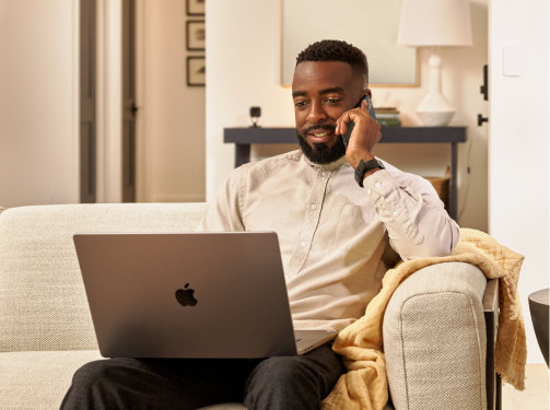 Man sitting in his living room with a laptop in his lap and talking on the phone
