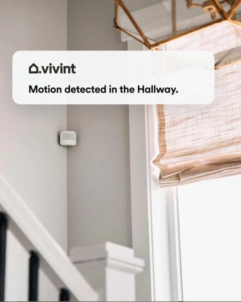 Vivint motion sensor on a wall next to a window, with a notification from the app saying "Motion detected in the hallway" hovered over