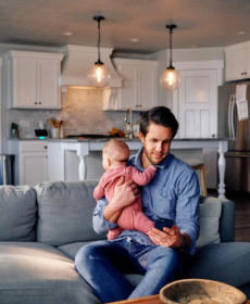 Dad holding baby while looking at phone on the couch and the kitchen in the background