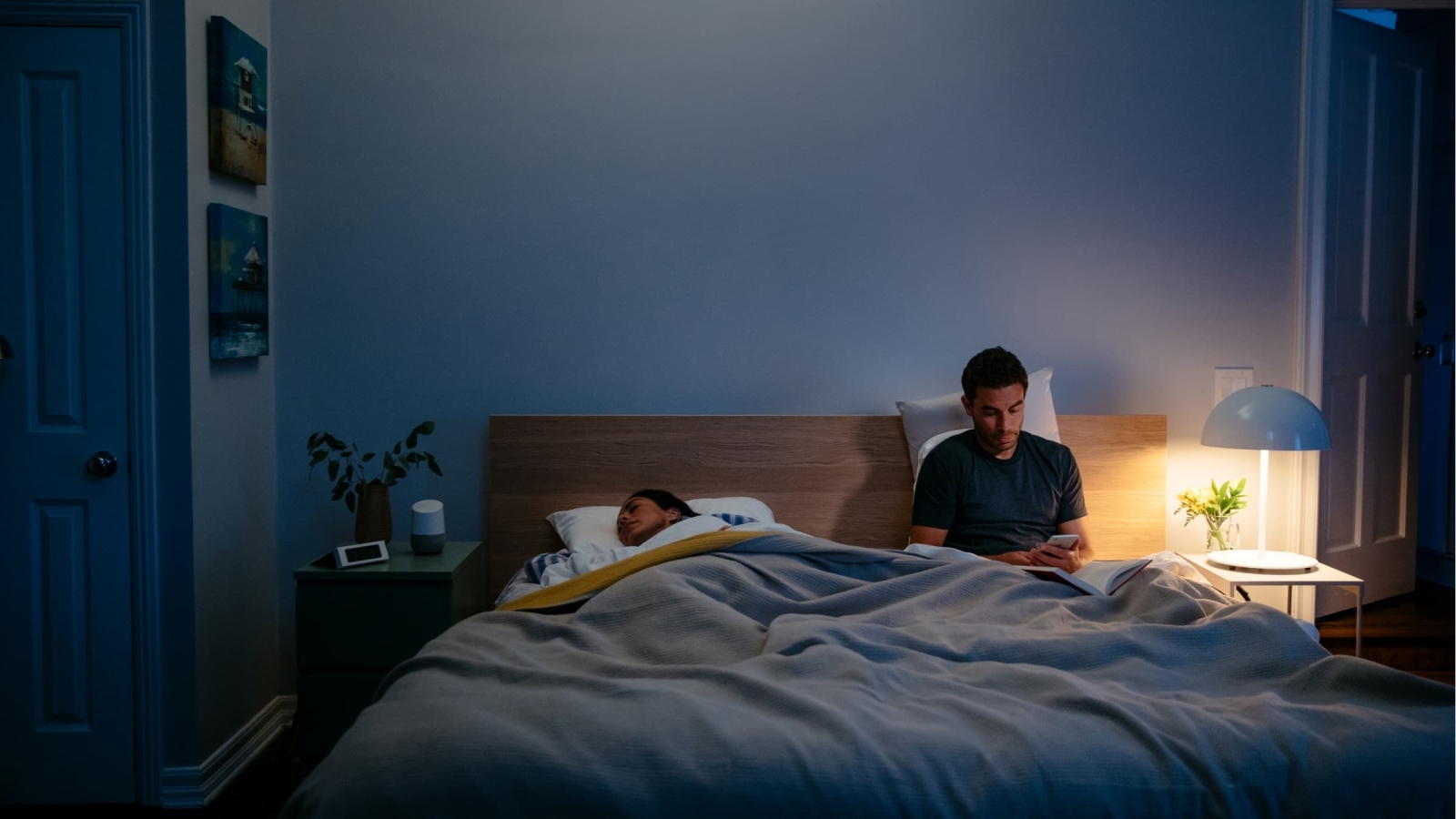 Man in bed next to partner on phone with an open book and door with a lit lamp on nightstand